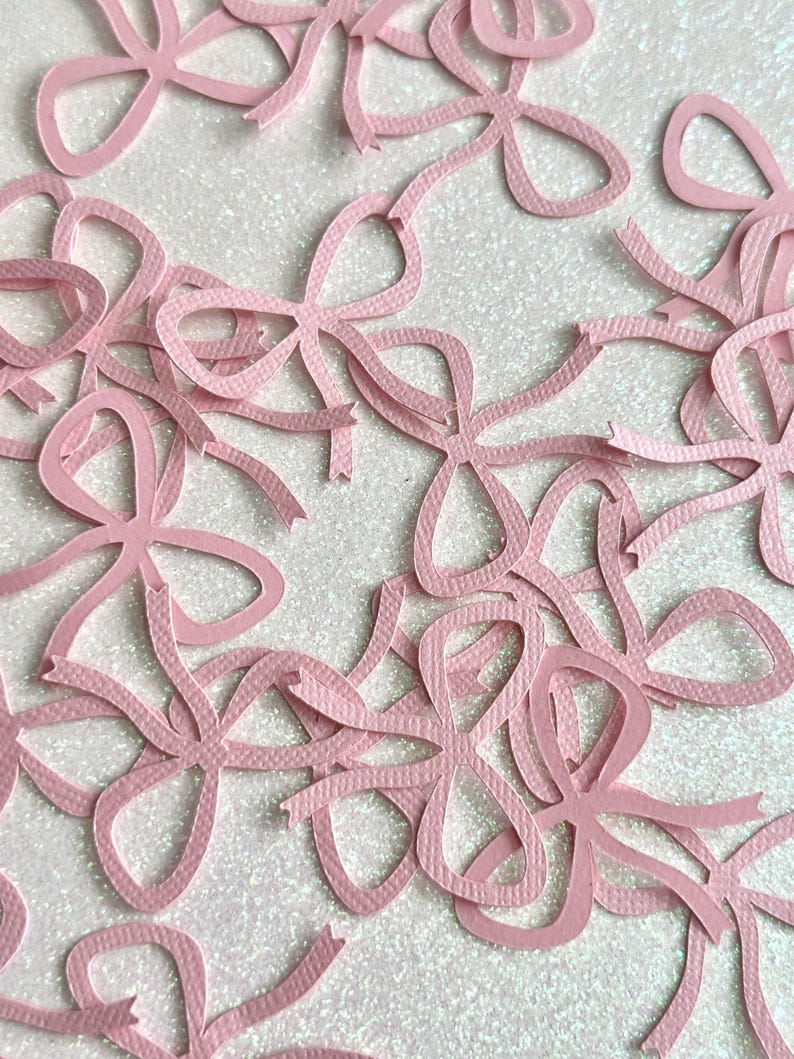 May include: A collection of light pink paper bows scattered across a white, glittery surface. The bows are cut from textured paper and feature a classic bow shape with long, flowing ribbon ends. Ideal for crafting and embellishments.