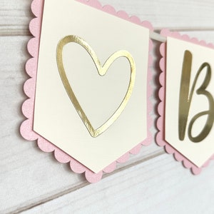Op de afbeelding: Een slinger met geschulpte roze randen en cr&egrave;mekleurige vlaggetjes. E&eacute;n vlaggetje heeft een gouden hartuitsparing, de andere de gouden letter "B". De slinger is geregen aan een wit lint, perfect voor een feest.