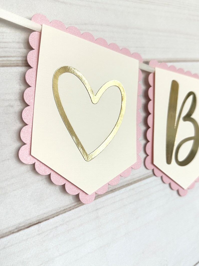 Op de afbeelding: Een slinger met geschulpte roze randen en cr&egrave;mekleurige vlaggetjes. E&eacute;n vlaggetje heeft een gouden hartuitsparing, de andere de gouden letter "B". De slinger is geregen aan een wit lint, perfect voor een feest.
