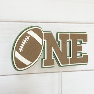 May include: A brown and green paper cake topper with the word "ONE" and a football. The football is brown with white stitching and a white outline.