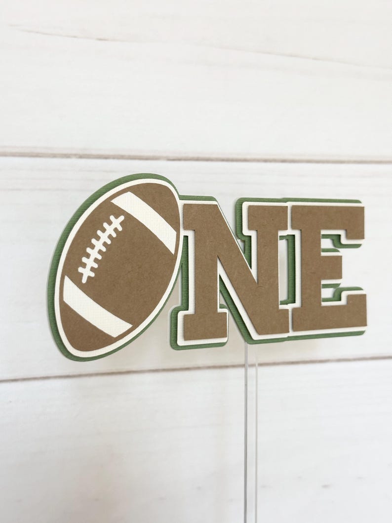 May include: A brown and green paper cake topper with the word "ONE" and a football. The football is brown with white stitching and a white outline.
