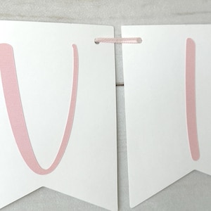 May include: White banner with pink letters and pink ribbon. The banner is made of white paper and has pink letters spelling out a word. The banner is strung together with pink ribbon.
