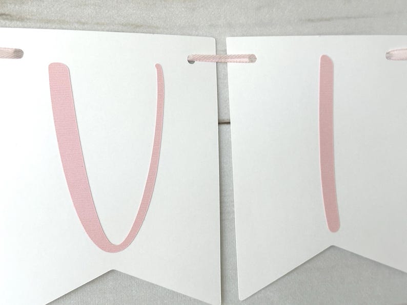 May include: White banner with pink letters and pink ribbon. The banner is made of white paper and has pink letters spelling out a word. The banner is strung together with pink ribbon.
