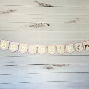 Op de afbeelding: Een slinger met de gouden tekst "Baby in Bloom". De slinger heeft een geschulpte roze rand en een witte achtergrond. Hij is geregen aan een wit touwtje en heeft aan elk uiteinde een hart.
