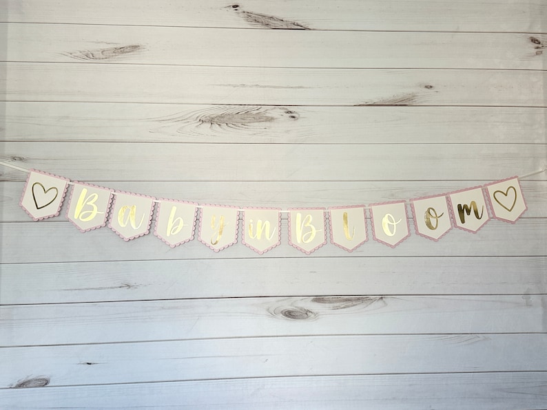 Op de afbeelding: Een slinger met de gouden tekst "Baby in Bloom". De slinger heeft een geschulpte roze rand en een witte achtergrond. Hij is geregen aan een wit touwtje en heeft aan elk uiteinde een hart.