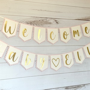 Op de afbeelding: Een slinger met de gouden tekst "Welcome Baby Ella". De slinger heeft off-white vlaggetjes met een roze geschulpte rand, geregen aan een wit lint. Perfect voor een babyshower of als decoratie.
