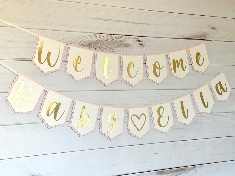 Op de afbeelding: Een slinger met de gouden tekst "Welcome Baby Ella". De slinger heeft off-white vlaggetjes met een roze geschulpte rand, geregen aan een wit lint. Perfect voor een babyshower of als decoratie.