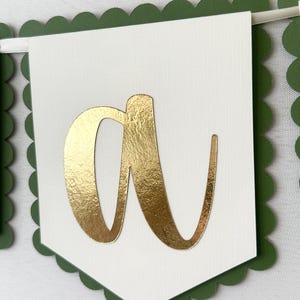 Op de afbeelding: Een slinger met een witte vlag met een gouden folie letter "a". De vlag heeft een geschulpte groene rand. De slinger is geregen aan een wit lint, ideaal voor feesten of gepersonaliseerde evenementen.