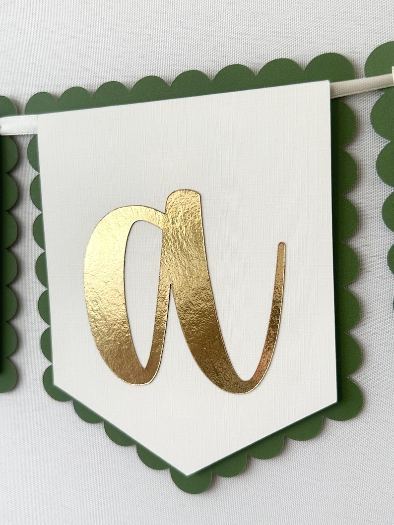 Op de afbeelding: Een slinger met een witte vlag met een gouden folie letter "a". De vlag heeft een geschulpte groene rand. De slinger is geregen aan een wit lint, ideaal voor feesten of gepersonaliseerde evenementen.