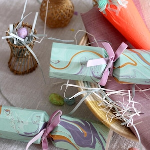 Può includere: Una natura morta a tema pasquale con cracker decorativi in carta marmorizzata, legati con nastri rosa. Una carota di carta, uova di Pasqua e piccoli cestini intrecciati creano un'atmosfera festosa.