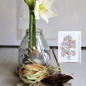 Könnte beinhalten: Ein Stillleben mit einer weißen Amaryllisblüte in einer transparenten, gesprenkelten Vase. Eine dekorative Karte mit floralem Design und eine getrocknete Proteablüte sind ebenfalls vorhanden. Das Arrangement steht auf einer strukturierten Oberfläche.