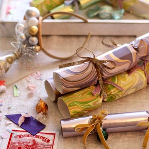 Puede incluir: Petardos festivos con diseños de papel veteado en morado, verde y plateado, atados con cintas doradas. Una botella de champán en miniatura y confeti se suman al tema festivo. Decoraciones de fiesta.