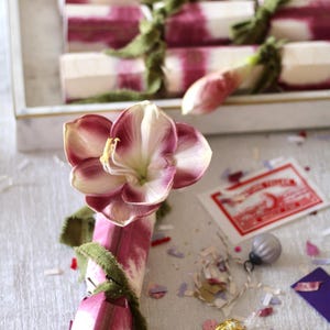 Op de afbeelding: Een feestelijke opstelling met decoratieve crackers met een roze en wit ontwerp, vastgebonden met groen lint. Een grote bloem met roze en witte bloemblaadjes is bevestigd aan een cracker. Confetti en een kleine versiering zijn verspreid.
