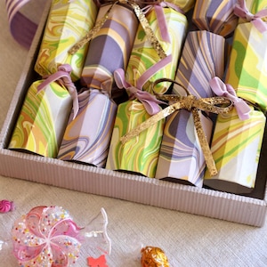 Puede incluir: Una bandeja de petardos de fiesta coloridos con papel veteado en tonos amarillos, verdes y morados, atados con cintas de terciopelo y doradas. La escena incluye caramelos dispersos y una gran piruleta rosa.