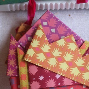 Op de afbeelding: Een verzameling kleurrijke papieren kronen met patronen in tinten oranje, roze en rood. De kronen zijn versierd met sterren en geometrische ontwerpen. Op de achtergrond een groene cracker met gouden sterren en rood fluwelen lint.