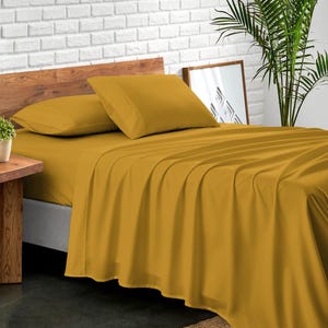May include: A mustard yellow fitted sheet on a bed with two matching pillowcases. The bed is made with a wooden headboard and a white brick wall in the background.