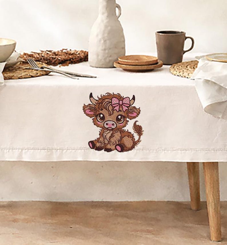 May include: Embroidered image of a Highland cow calf with a pink bow. The calf is brown with pink accents on its ears and hooves. The image is on a white tablecloth, with a neutral background.