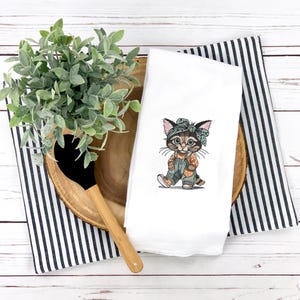 Puede incluir: Toalla de cocina blanca con un gatito de dibujos animados bordado que lleva un mono y una bandana. El gatito est&aacute; representado en tonos marrones, naranjas y azules. La toalla est&aacute; colocada sobre una bandeja de madera con una toalla a rayas blancas y negras y una cuchara de madera.