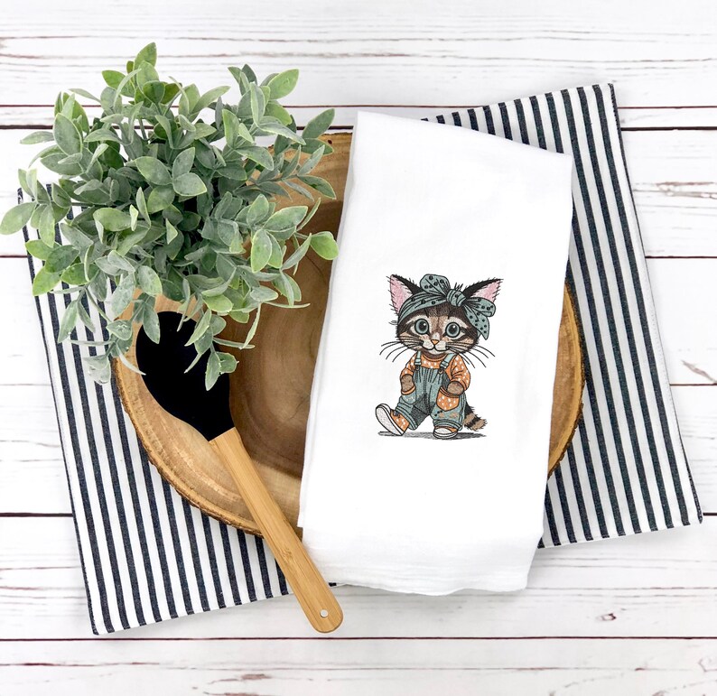 Puede incluir: Toalla de cocina blanca con un gatito de dibujos animados bordado que lleva un mono y una bandana. El gatito est&aacute; representado en tonos marrones, naranjas y azules. La toalla est&aacute; colocada sobre una bandeja de madera con una toalla a rayas blancas y negras y una cuchara de madera.