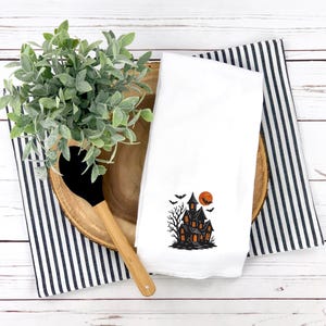 May include: White kitchen towel with a black and orange haunted house design, bats, and a full moon. The towel is folded and placed on a wooden tray with a black and white striped cloth and a wooden spoon.