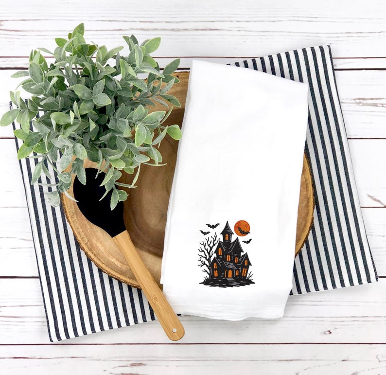 May include: White kitchen towel with a black and orange haunted house design, bats, and a full moon. The towel is folded and placed on a wooden tray with a black and white striped cloth and a wooden spoon.