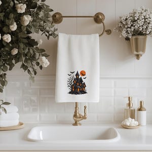 May include: White Halloween-themed hand towel with a spooky haunted house design. The black and orange graphic features a haunted house, bats, and a full moon. The towel is hanging on a gold towel rack in a bathroom setting.