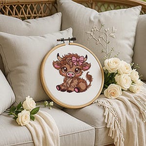 May include: A cross-stitch embroidery hoop showing a cartoon Highland cow with a pink bow. The cow is brown and pink, with large eyes. The hoop is on a beige sofa with white roses and pillows.