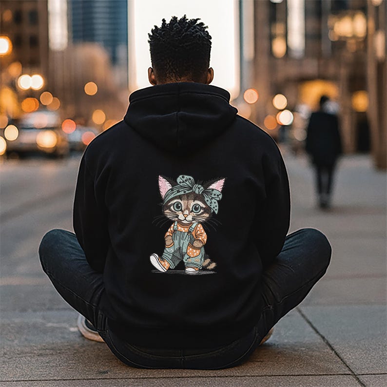 Puede incluir: Sudadera con capucha negra con un dise&ntilde;o de gato de dibujos animados en la espalda. El gato lleva un mono y una bandana. La persona de la imagen lleva vaqueros azul oscuro y est&aacute; sentada sobre una superficie de hormig&oacute;n. El fondo est&aacute; borroso.