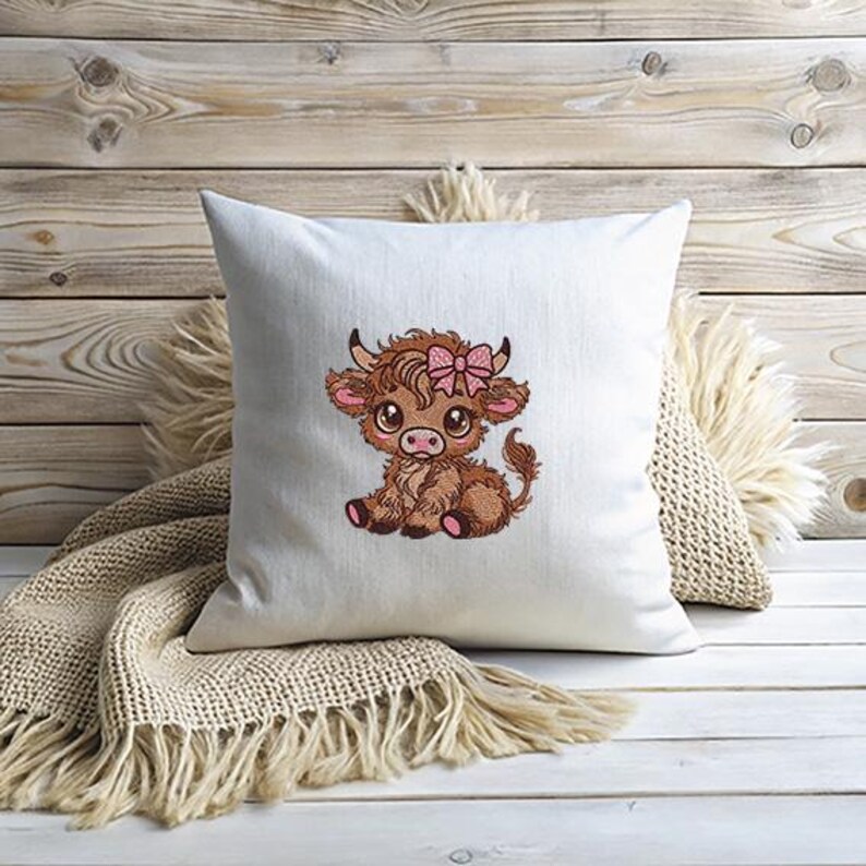 May include: A white square cushion featuring an embroidered cartoon Highland cow. The cow is brown with a pink bow, in a seated position. The cushion rests on a light surface with a fringed throw.