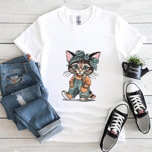 Puede incluir: Camiseta blanca con un gato de dibujos animados con un peto y una bandana. El gato es marr&oacute;n, canela y blanco con un peto azul y una bandana a juego. Unos vaqueros azules y zapatillas deportivas blancas y negras est&aacute;n al lado de la camiseta.