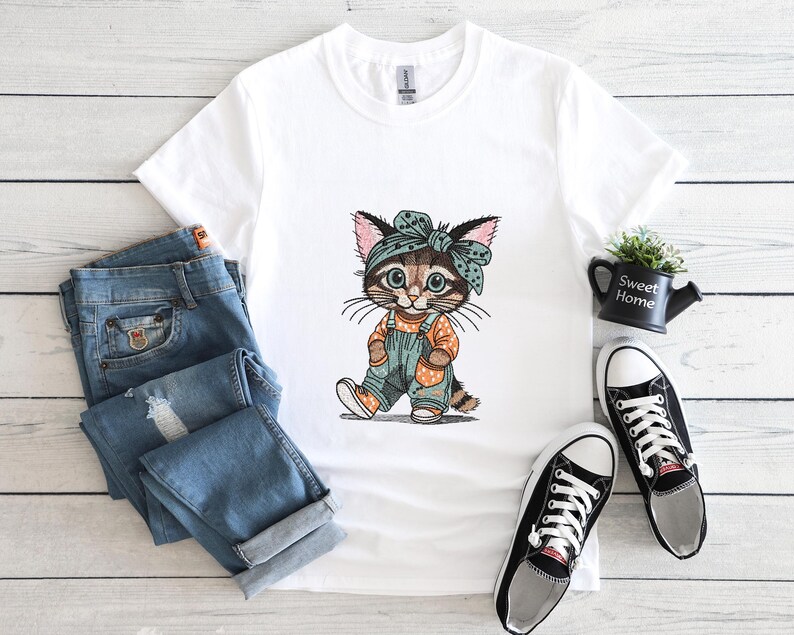 Puede incluir: Camiseta blanca con un gato de dibujos animados con un peto y una bandana. El gato es marr&oacute;n, canela y blanco con un peto azul y una bandana a juego. Unos vaqueros azules y zapatillas deportivas blancas y negras est&aacute;n al lado de la camiseta.