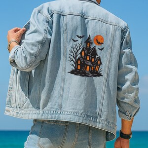 May include: Light blue denim jacket featuring a Halloween-themed graphic on the back. The graphic includes a haunted house, bats, and a full orange moon. The jacket has a classic design with button closures and pockets.