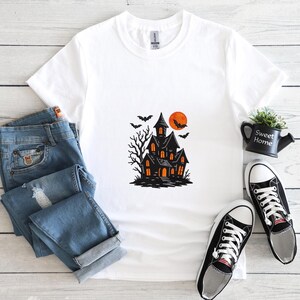 May include: White t-shirt featuring a black haunted house graphic with an orange moon and bats. The design includes a spooky tree and glowing windows. The shirt is paired with blue jeans and black and white sneakers, creating a Halloween theme.