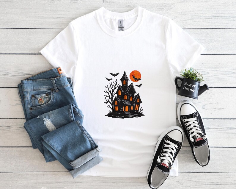 May include: White t-shirt featuring a black haunted house graphic with an orange moon and bats. The design includes a spooky tree and glowing windows. The shirt is paired with blue jeans and black and white sneakers, creating a Halloween theme.