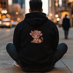 May include: Black hoodie with a cartoon Highland cow illustration on the back. The cow is brown with a pink bow. The hoodie is worn by a person sitting on the ground.
