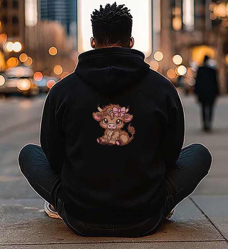 May include: Black hoodie with a cartoon Highland cow illustration on the back. The cow is brown with a pink bow. The hoodie is worn by a person sitting on the ground.