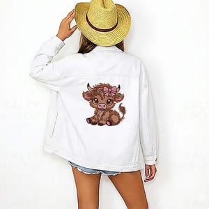 May include: White denim jacket with a cartoon Highland cow and a pink bow. The jacket has a classic design with a button-up front, pockets, and a collar. The cow graphic is embroidered on the back.