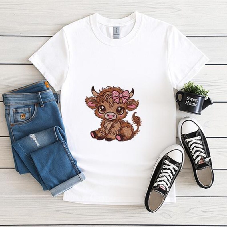 May include: White t-shirt with an embroidered cartoon Highland cow wearing a pink bow. The shirt is styled with blue jeans and black canvas trainers. A small black watering can with the words "Sweet Home" is also visible.