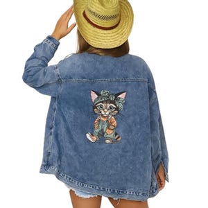 Puede incluir: Chaqueta vaquera azul claro con un dibujo de gato de dibujos animados en la espalda. El gato lleva un peto y una diadema. La chaqueta tiene un cuello cl&aacute;sico y cierre de botones.