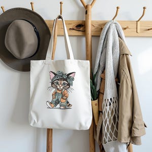 Puede incluir: Un bolso tote blanco con una ilustraci&oacute;n de gato de dibujos animados cuelga de un perchero de madera. El gato lleva un peto y una diadema. Un sombrero marr&oacute;n y un abrigo beige tambi&eacute;n est&aacute;n en el perchero.