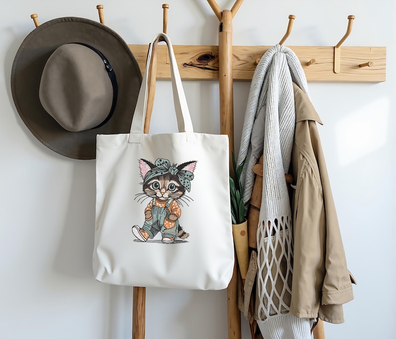 Puede incluir: Un bolso tote blanco con una ilustraci&oacute;n de gato de dibujos animados cuelga de un perchero de madera. El gato lleva un peto y una diadema. Un sombrero marr&oacute;n y un abrigo beige tambi&eacute;n est&aacute;n en el perchero.
