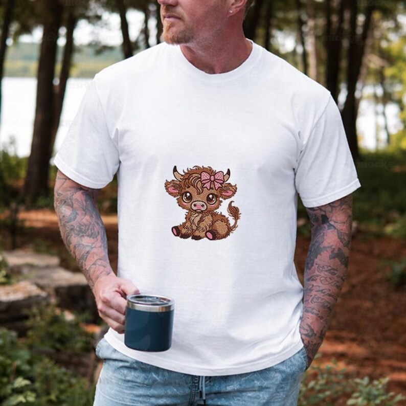 May include: White t-shirt with a cartoon Highland cow calf illustration. The calf is brown and tan, wearing a pink bow, and sitting down. The t-shirt has short sleeves and is plain. The image is set against a natural background.