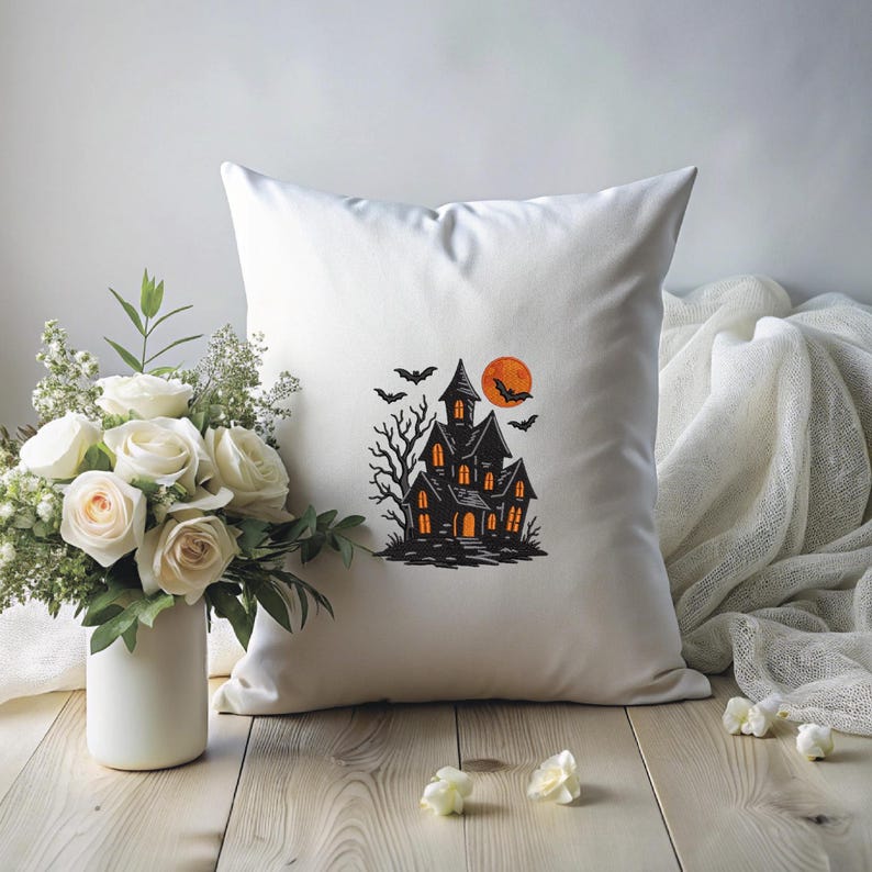 May include: White square pillow featuring a black haunted house design with an orange moon and bats. The pillow is on a wooden surface with a bouquet of white roses and a white cloth in the background. Halloween decor.
