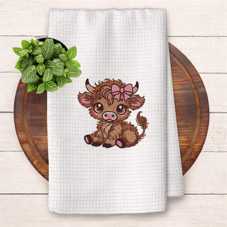 May include: White waffle-weave kitchen towel with an embroidered brown Highland cow wearing a pink bow. The cow has large eyes and is sitting down. The towel is on a wooden chopping board with a small green plant in a black pot.