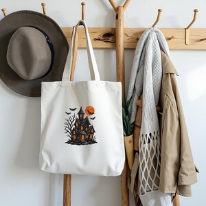 May include: A white tote bag with a Halloween-themed design featuring a haunted house, bats, and a full orange moon. The bag is hanging on a wooden coat rack with a hat and other clothing items.