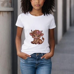 May include: White t-shirt with a cartoon Highland cow design, complete with a pink bow. The cow is brown with black horns and is sitting. The shirt is worn with blue jeans. The model is standing in front of a grey wall.