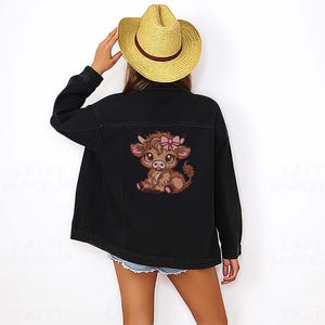 May include: Black denim jacket with an embroidered cartoon Highland cow wearing a pink bow. The jacket has a classic collar, button-up front, and long sleeves. The cow graphic is on the back. The model is wearing a straw hat and denim shorts.
