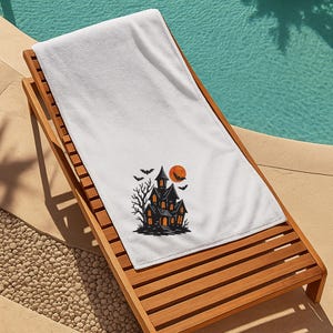 May include: White beach towel with a Halloween-themed design. The design features a black haunted house with orange lit windows, a full orange moon, bats, and a bare tree. The towel is lying on a wooden lounge chair.