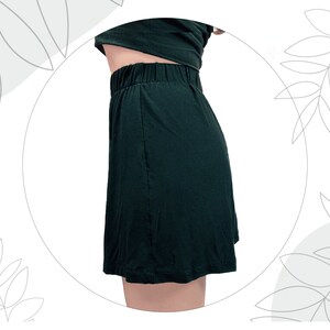 May include: A dark green skirt with an elasticized waistband and a simple, straight-cut silhouette. The garment is shown from a side profile view, featuring a smooth, solid-colored fabric that falls to a hemline above the knee.