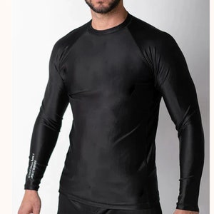 May include: A solid black long-sleeve compression shirt with a crew neckline and raglan sleeves. The fabric has a smooth, slightly reflective finish. A small white text graphic is printed vertically on the lower portion of the right sleeve.